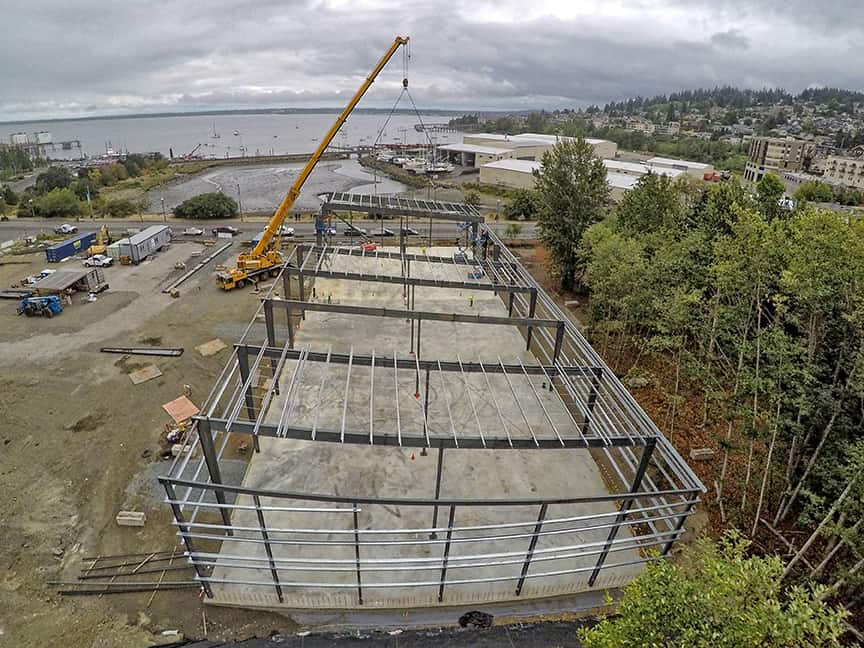 Steel structural frame mid-erection at the 700 Harris commercial site — yellow crane setting beams above the Bellingham Bay waterfront.